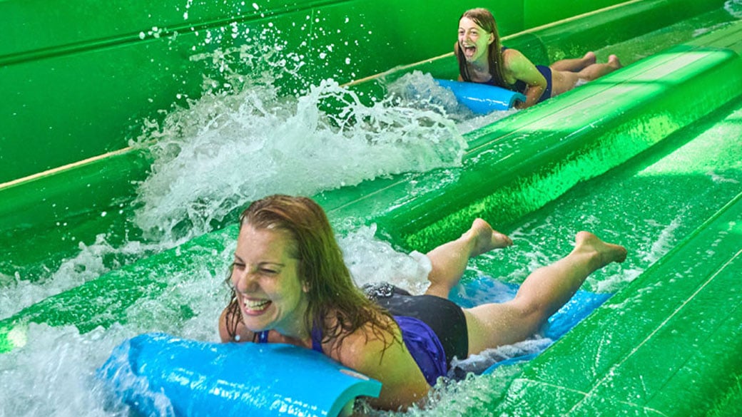 Two girls sliding down a green water slide inside the lobby of Great Wolf Lodge 